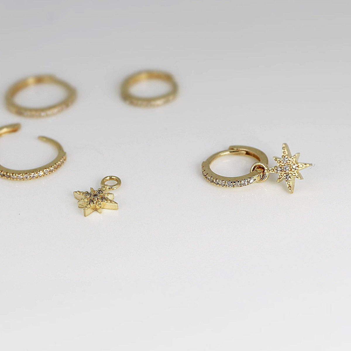 Gold jewelry pieces including a pave huggie hoop, a huggie with a star charm, a loose star charm, and two plain gold bands on a white surface.