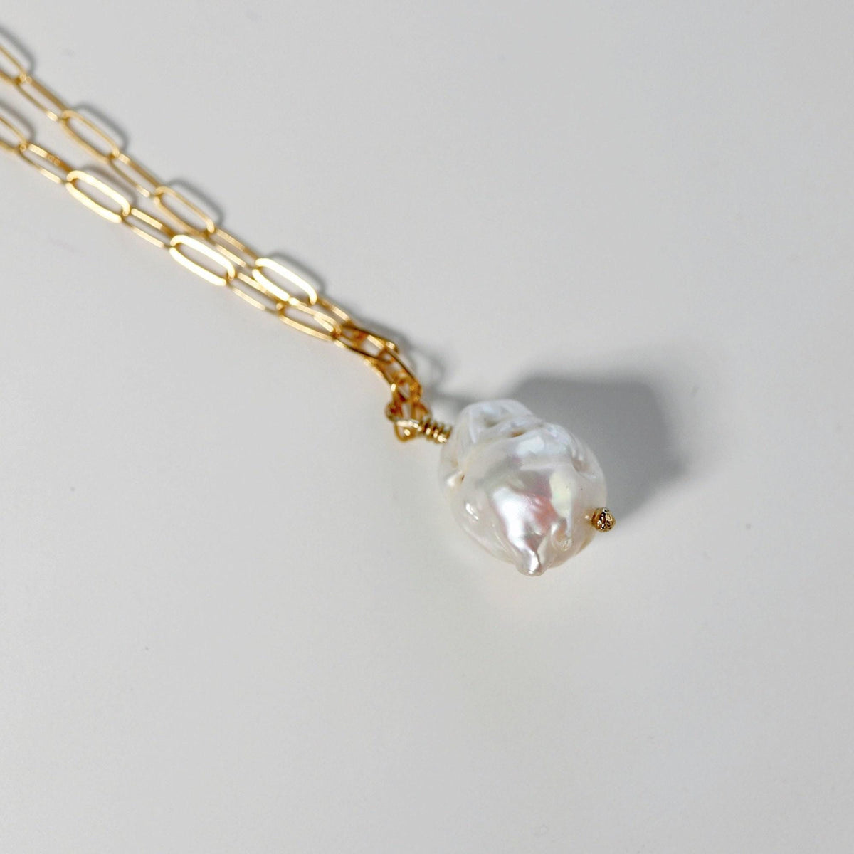 Angled close-up of a gold paperclip chain and white baroque pearl pendant resting on a light gray surface.