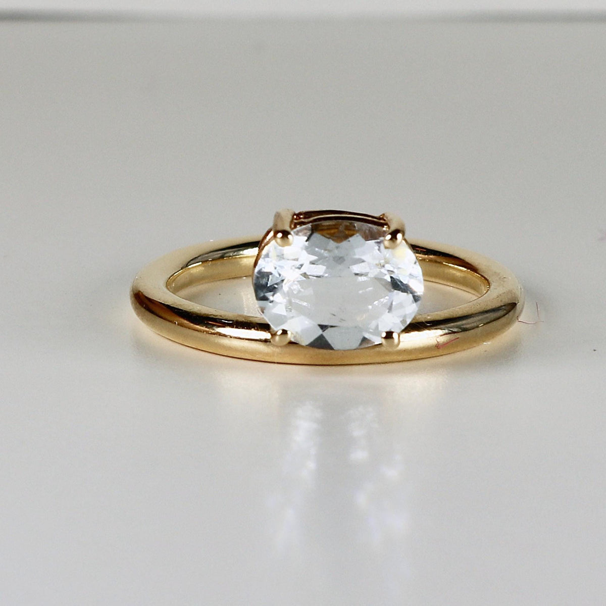 A gold ring with a horizontally set, faceted clear oval gemstone rests on a white surface, viewed from the side.
