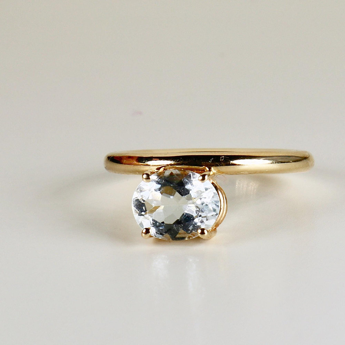 A gold ring featuring a clear faceted oval gemstone set vertically rests on a neutral surface.