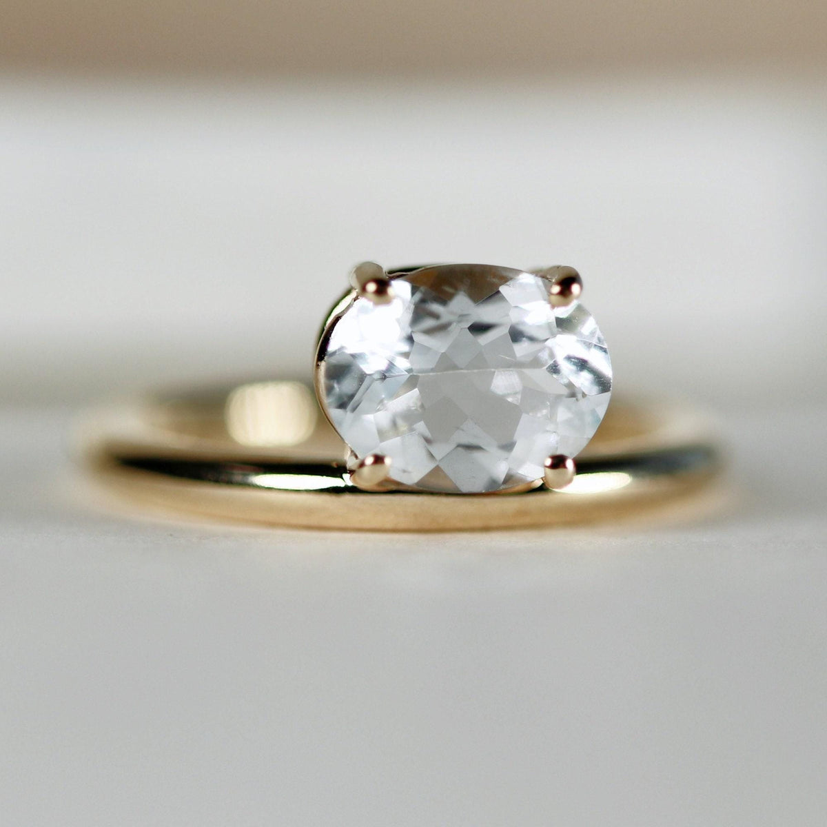 A gold ring with an east-west set, faceted clear oval gemstone rests on a light background.
