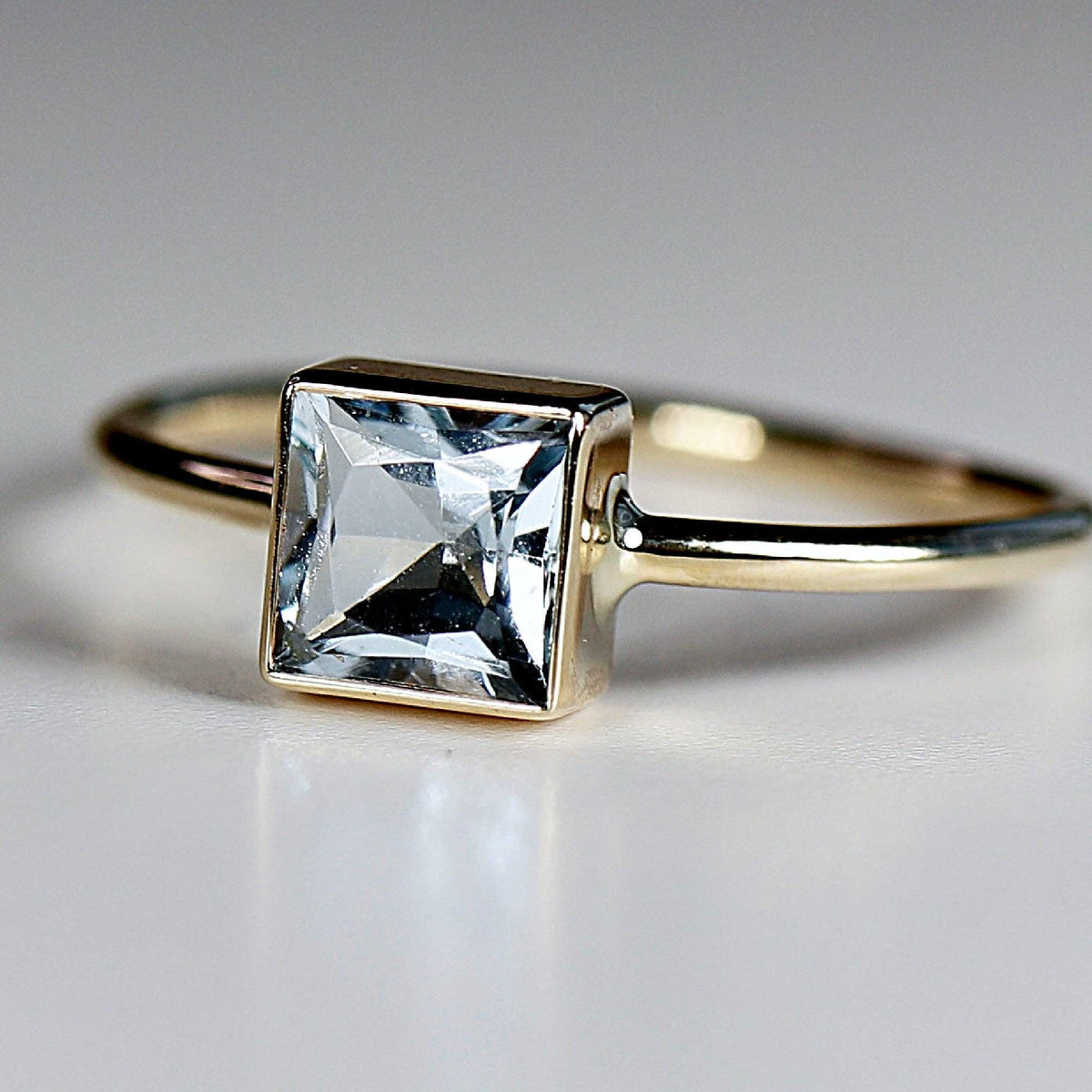 Close up angled view of a gold ring featuring a bezel set square light blue gemstone on a white surface.