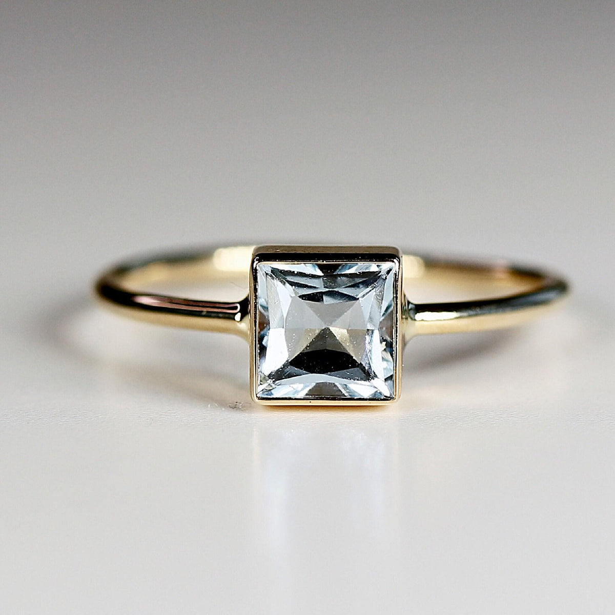Front facing close up view of a gold ring with a bezel set square faceted light blue stone.