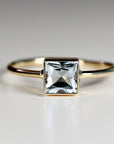 Front facing close up view of a gold ring with a bezel set square faceted light blue stone.
