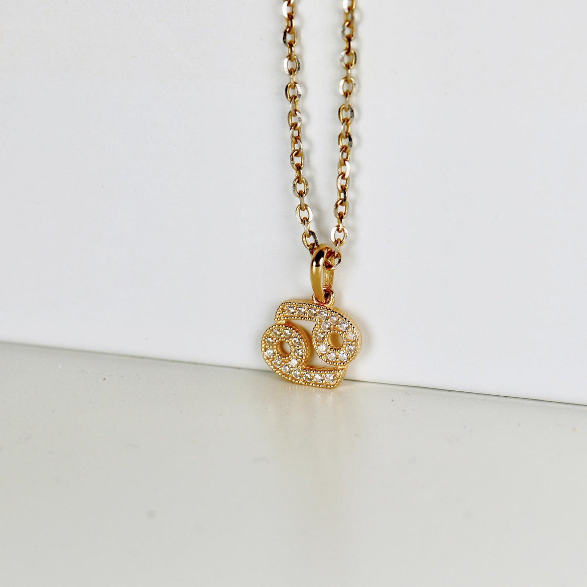 A gold chain and a Cancer astrological pendant with inlaid white stones rest on a white display surface.