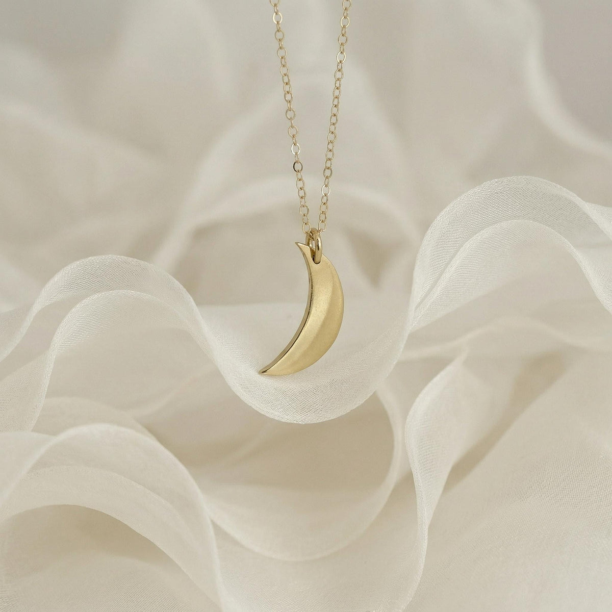 A gold crescent moon pendant necklace displayed on soft, draped white fabric.