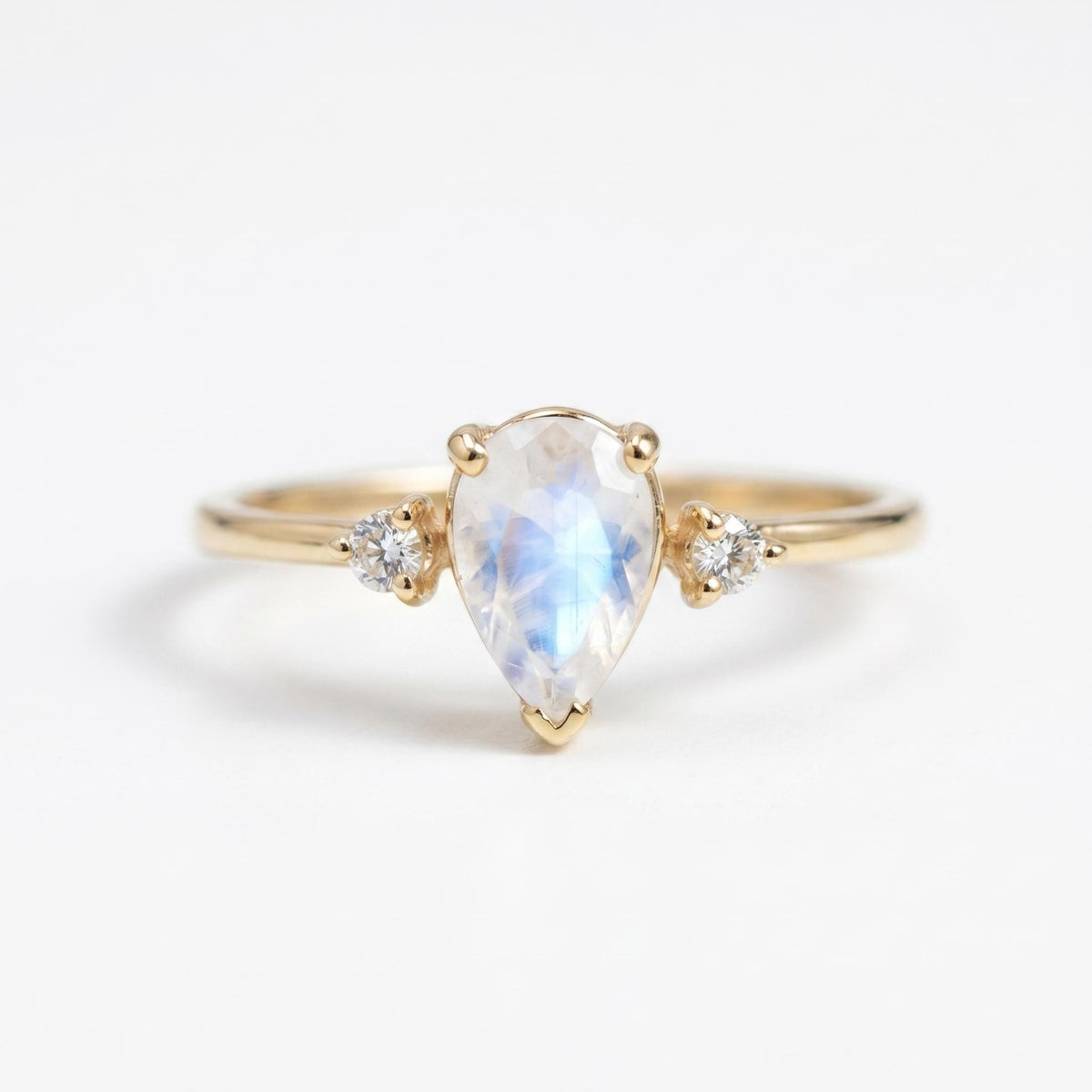 A gold ring centered with a pear-shaped rainbow moonstone and flanked by two small round diamonds against a white background.