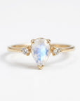 A gold ring centered with a pear-shaped rainbow moonstone and flanked by two small round diamonds against a white background.