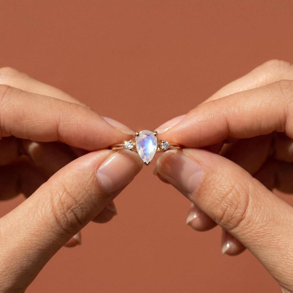 Two hands hold up the gold pear moonstone and diamond ring against a terracotta-colored background.