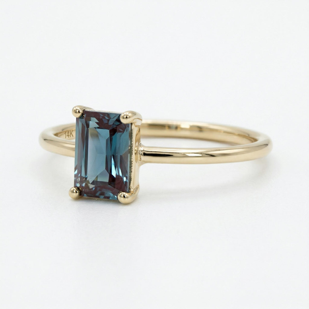 A yellow gold ring with an emerald-cut blue-green alexandrite gemstone shown angled against a plain white background.
