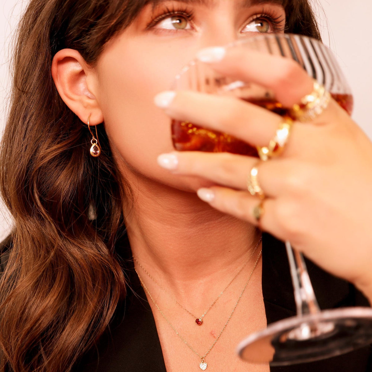 A woman wearing gold drop earrings with pink teardrop stones, two gold necklaces, and gold rings while holding a glass.