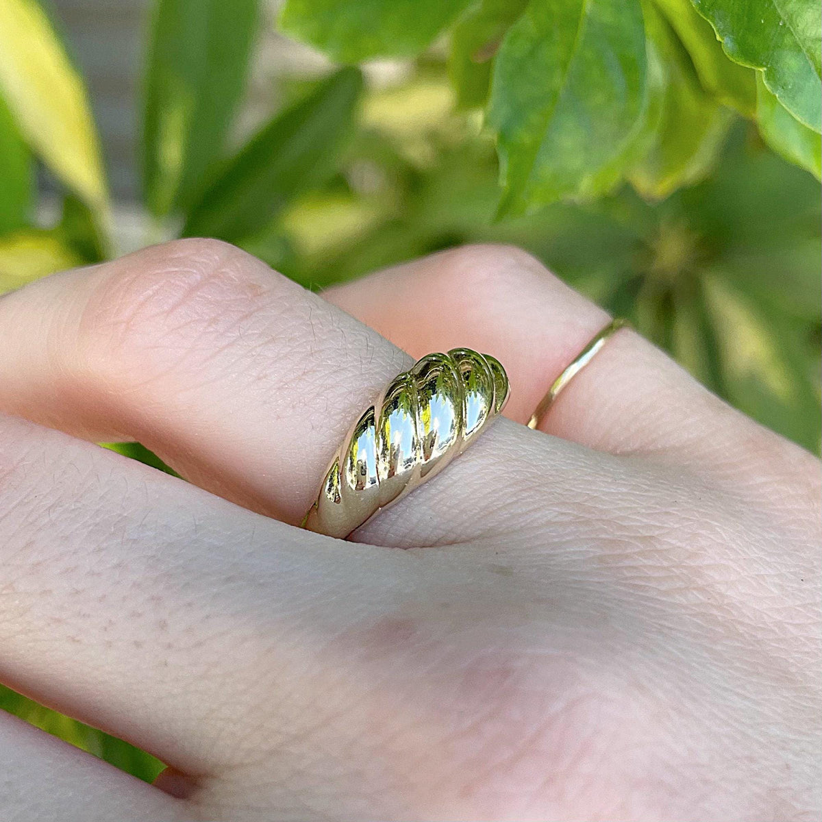 An outdoor macro shot of a gold croissant ring worn on a finger with green foliage in the background.