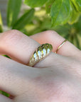 An outdoor macro shot of a gold croissant ring worn on a finger with green foliage in the background.