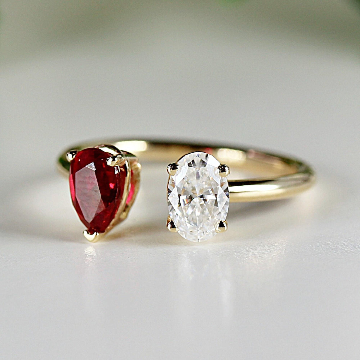 A gold bypass ring featuring a pear-cut red stone on the left and an oval-cut white stone on the right, resting on a white surface.