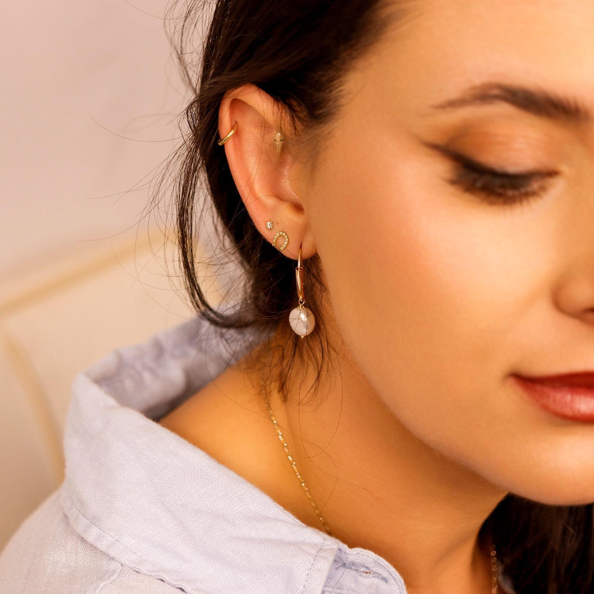 Profile shot of a woman wearing the gold pearl dangle earring and multiple small stud earrings, dressed in a light blue button-down shirt.