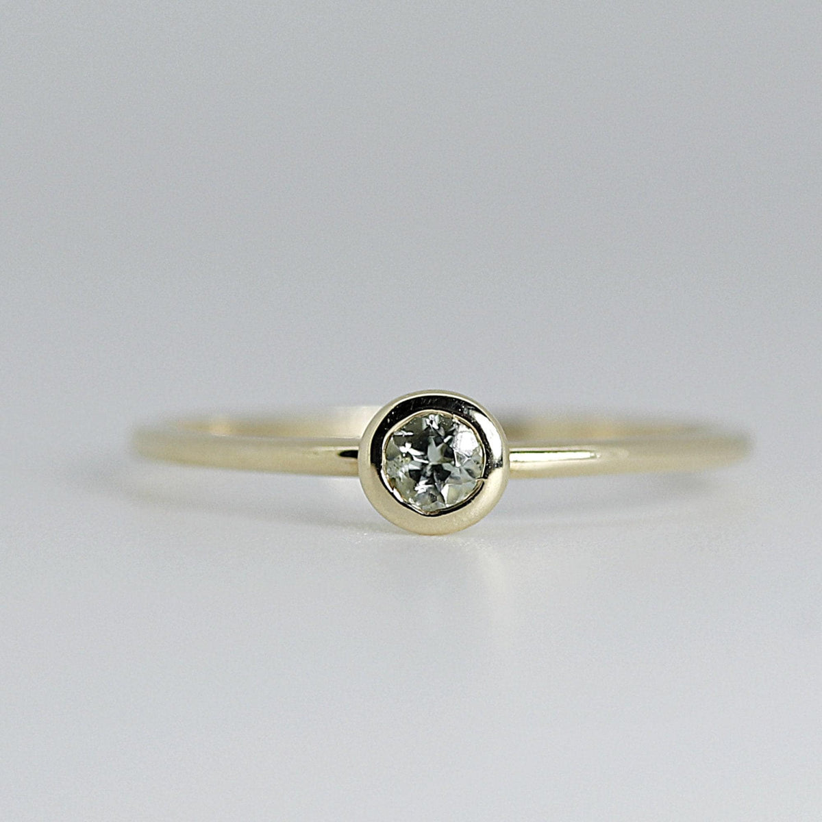 Front-facing, close-up view of a thin gold ring with a round, bezel-set light green gemstone.