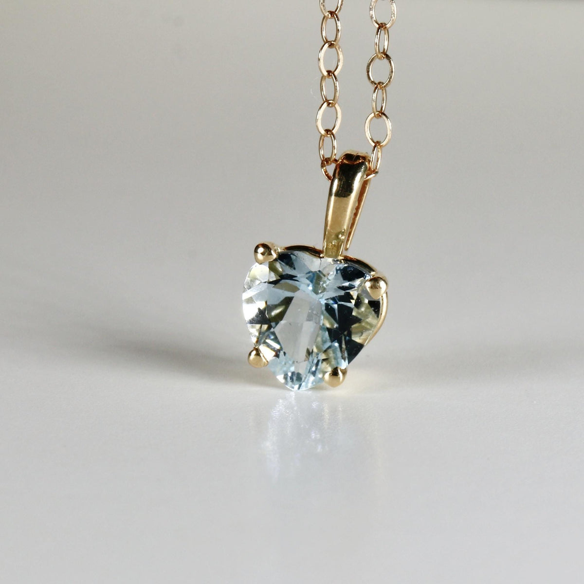 A heart-shaped pale blue gemstone pendant on a gold chain, viewed closely against a white backdrop.