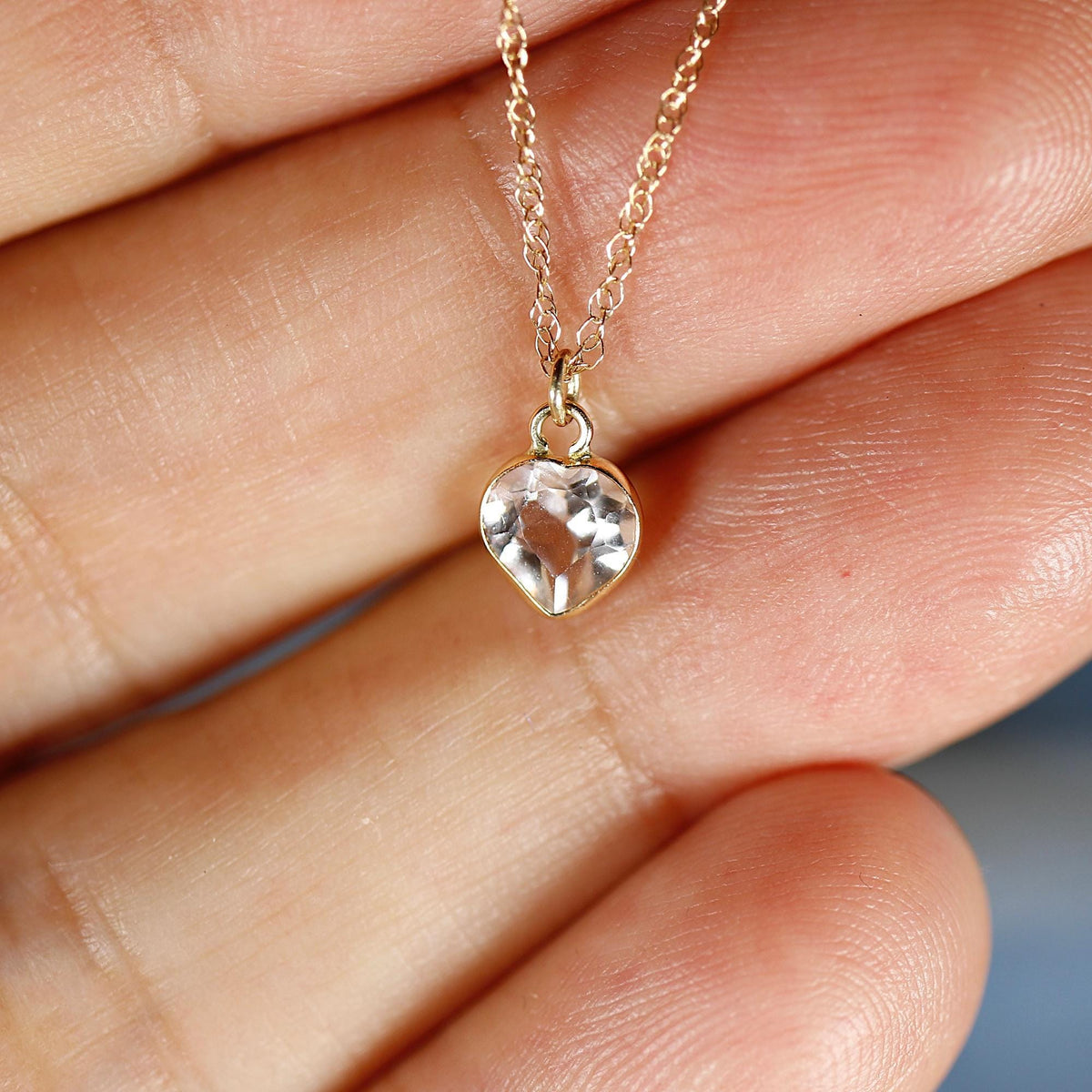 A gold chain necklace with a small faceted heart-shaped clear gemstone pendant rests on a person&amp;