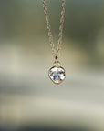 A gold chain necklace with a heart-shaped clear gemstone pendant hangs against a soft-focus outdoor background.