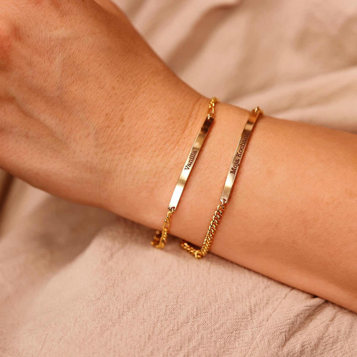 Two gold bar bracelets worn on a wrist against a neutral fabric background.