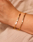 Two gold bar bracelets worn on a wrist against a neutral fabric background.