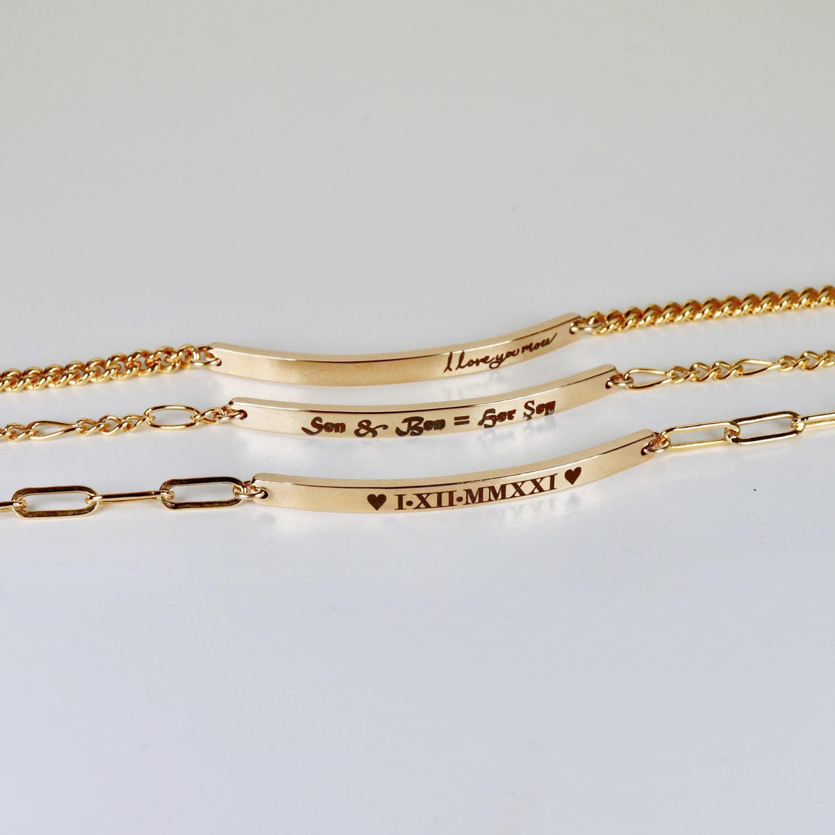 Three gold bracelets arranged vertically on a white surface, each featuring a personalized engraved bar pendant.