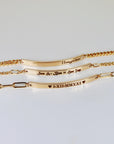Three gold bracelets arranged vertically on a white surface, each featuring a personalized engraved bar pendant.