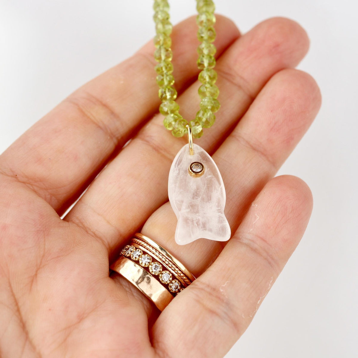 A hand holding a green peridot beaded necklace with a white stone fish pendant and several rings.