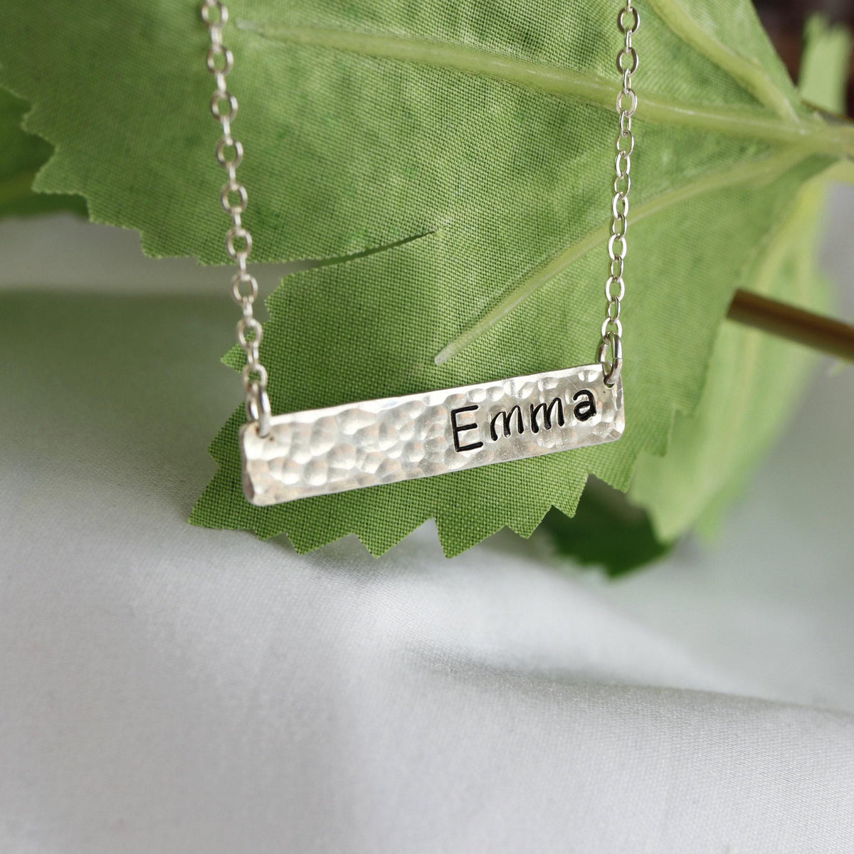 A silver bar necklace rests against a green leaf and white fabric, stamped with the name Emma.