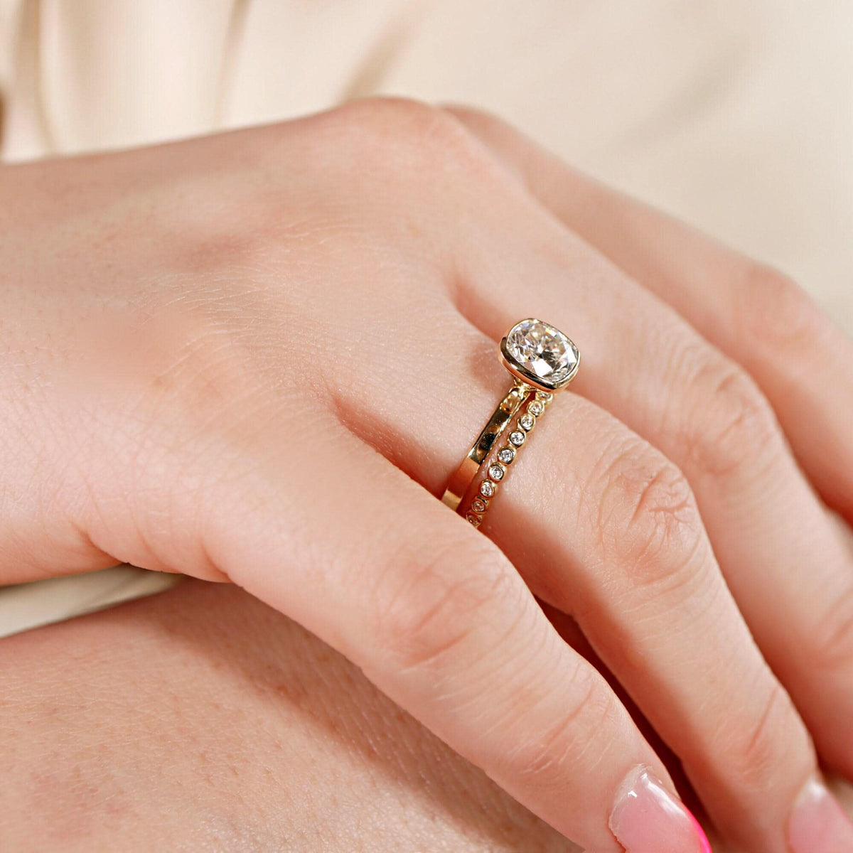A gold bezel-set cushion cut ring paired with a matching pavé diamond band worn on a finger.