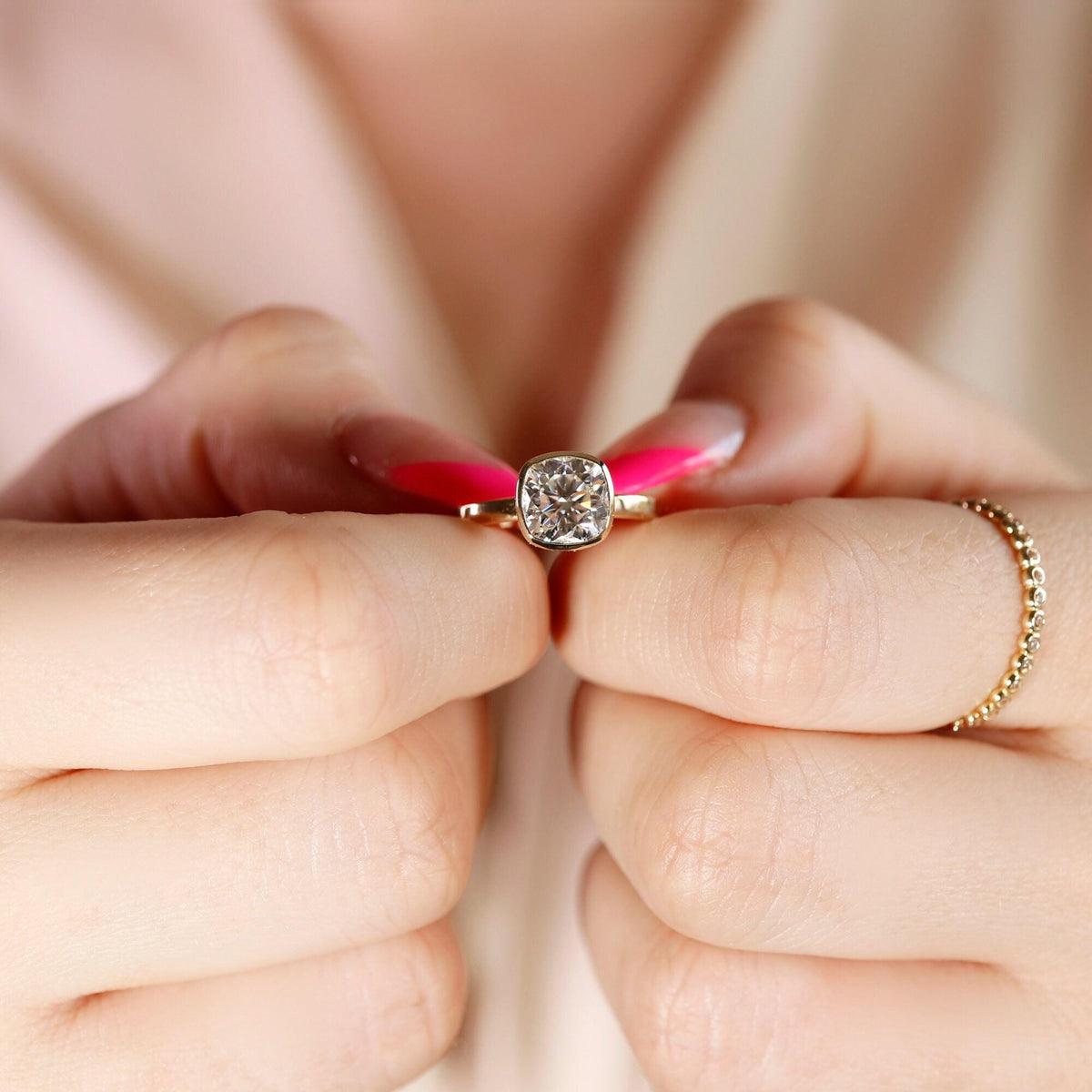 Hands holding a gold bezel-set cushion cut ring with a separate beaded band on the right finger.