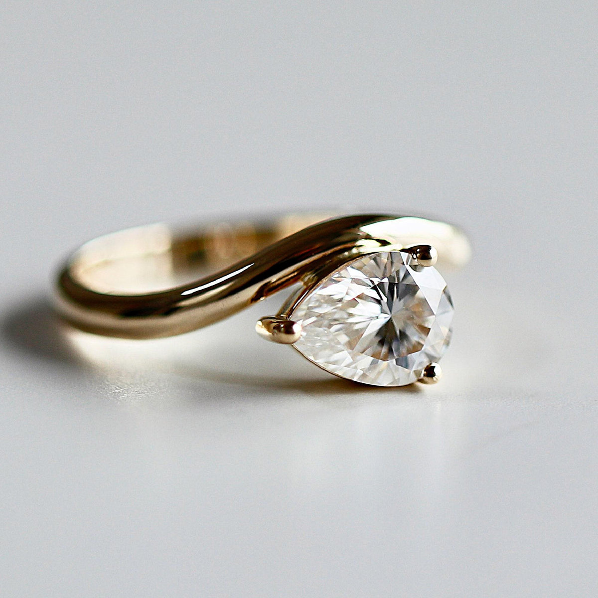 A studio shot of the gold pear-shaped solitaire ring angled on a light gray surface.