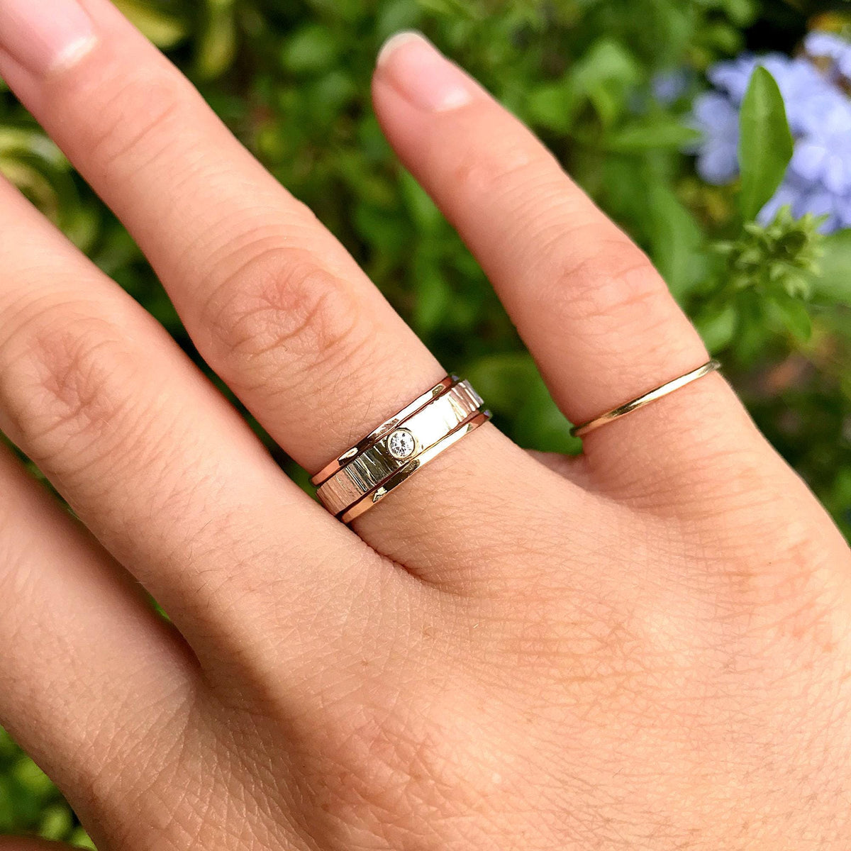 A hand wearing the stacked ring set on the ring finger and a thin gold band on the pinky finger, with blurred green foliage in the background.